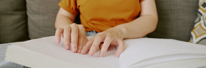 Website header of unrecognizable woman with visual impairment reading Braille book on sofa in...