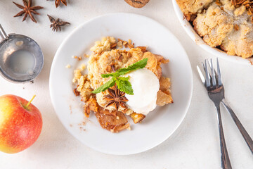 Apple Crumble Dessert, crisp cobbler with Ice Cream, crumble pie with crunchy dough topping in baking dish and plate, with ice cream scoop on white kitchen table