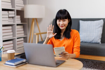 asian woman is sitting on a white couch in a modern home. Her is smiling and looking at her laptop freelance business online concept.

