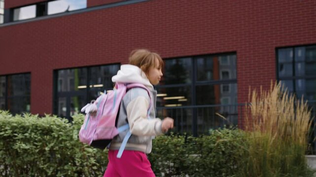 Young girl, around 7-9 years old, is joyfully skipping towards school. She wears a grey and white varsity jacket, bright pink pants, and a pink backpack with blue straps.