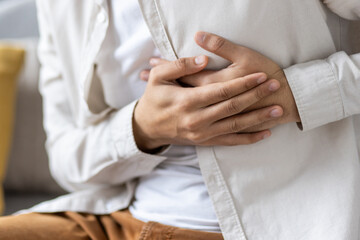 Person experiencing stomach pain with hands clutching abdomen, wearing casual clothing while sitting indoors