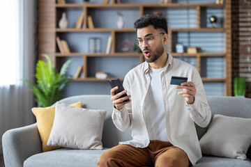 Shocked man holding smartphone and credit card sitting on sofa revealing online payment error