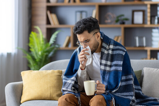 Young man sitting on sofa covered with blanket, drinking hot beverage and blowing nose, feeling unwell and cold