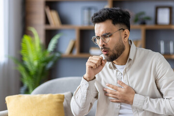 Man experiencing cough and chest discomfort at home, wearing glasses and casual clothing,...