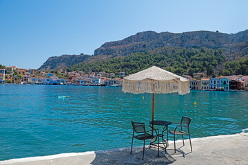 Sun umbrella, table and chairs on the beach. Sea, colourful houses and mountains in the background.