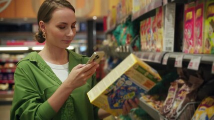 Young woman is shopping in a grocery store and checking her shopping list on her smartphone app. She is holding a box of cereal in her hand