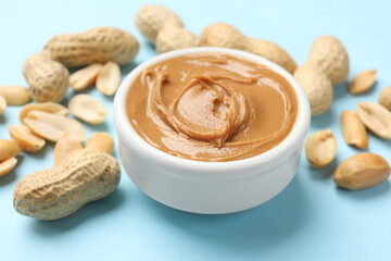 Tasty peanut butter in bowl and groundnuts on light blue background, closeup