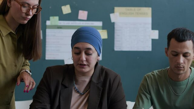 Medium shot of young Caucasian female teacher helping intercultural group of students sitting at desk and studying English grammar in classroom