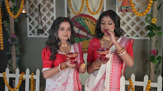 Two Indian women, draped in traditional sarees and adorned with gold jewellery, holding dhunachi in hands in front of a beautifully decorated house