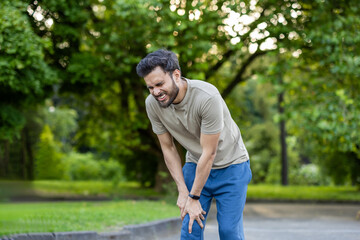 Man experiencing pain during exercise in park while engaged in outdoor fitness routine surrounded...