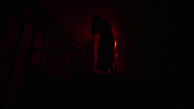 Creepy ghost woman in white dress and scary makeup standing alone with bended body on staircase in dark haunted house. Halloween horror advertisement concept.