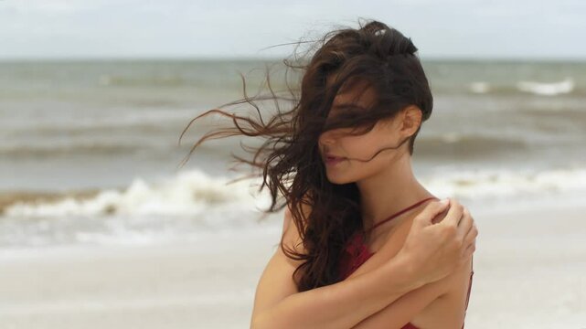 Pensive Walking Woman in Red Dress Embracing Herself on Windy Beach