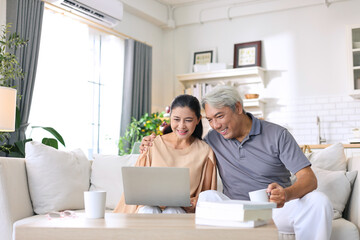 Asian senior woman happily watching movie on laptop with her elderly husband on sofa at home.
