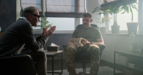 Caucasian military man in uniform sits in chair, talks to psychiatrist and discusses progress after treatment or psychotherapy. Senior psychotherapist smiles, listens to male soldier, asks questions.