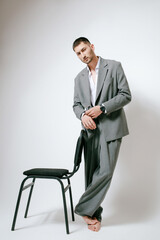 Man in grey costume stands near the chair, barefoot. In studio, fashion