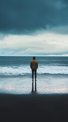 A solitary figure stands on a beach facing the ocean, with waves crashing gently onto the shore. The person wears a yellow jacket and appears to be in deep thought or contemplation, gazing at the hori