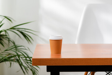 A disposable coffee cup on a wooden table beside a leafy plant