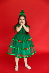 Surprised happy little girl in a green festive carnival costume of little Christmas tree on a red background. Winter holidays concept. Sale and advertising, copy space