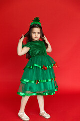 A little girl in a green festive carnival costume of little Christmas tree on a red background. Winter holidays concept. Sale and advertising, copy space