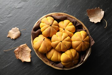 Wicker basket with tasty pumpkin shaped buns and dry leaves on dark textured table, flat lay