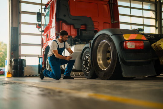A experienced mechanic in blue overalls kneels beside a large truck, inspecting the tire while holding a tablet.