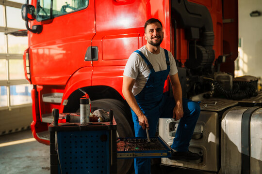 A smiling mechanic in blue work suit poses next to a red truck in a garage.
