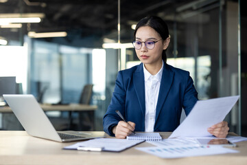 Businesswoman studies financial documents in office. She focuses on paperwork, analyzing data while...