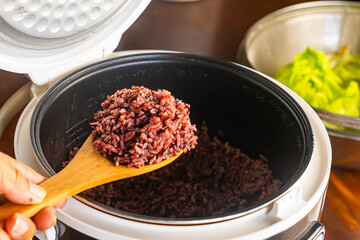 High angle view of hot steamed riceberry, rice berry in wooden spoon scooped from opened rice cooker with green vegetables on wooden table.