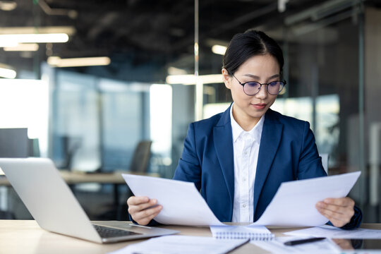 Professional woman in business attire analyzing important documents at desk. Focused, serious expression indicates careful consideration of financial data. Modern office environment