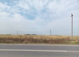 Asphalt road, then - field with mostly yellow grass, also - poles with electrical wires, blue sky with clouds. Outdoor. Landscape.
