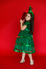 Happy little girl in a festive carnival costume of little Christmas tree with Christmas gift on red background. A little girl holds a gift on a red isolated background.