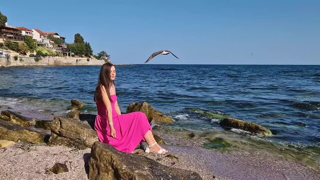 Beautiful young woman wearing pink dress, sitting on a rock at the beach watching the sights of the Nessebar old town in Bulgaria. Beautiful ancient houses on the coast. Summer vacation travel scene
