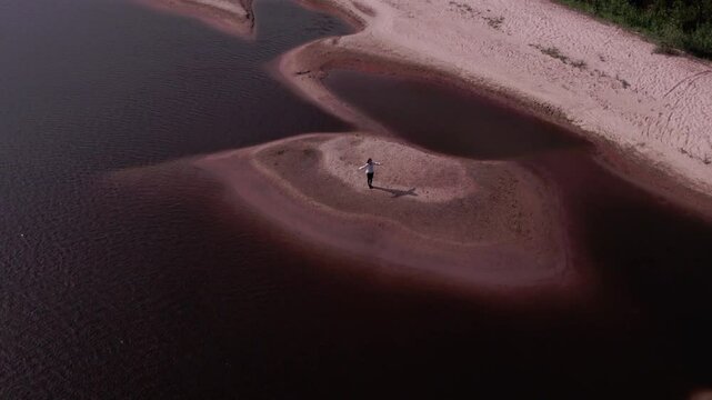 Drone footage of a lone man standing on a sandy island surrounded by water with his arms raised, casting a shadow on the island, the water glistening in the sun