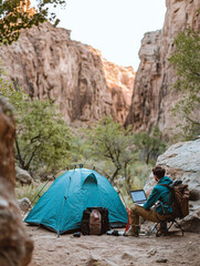 Fototapeta premium A man sits in front of a blue tent, laptop, and backpack