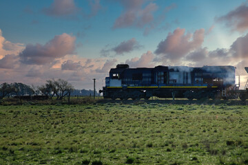 Old diesel locomotive steaming through a rural landscape at sunset