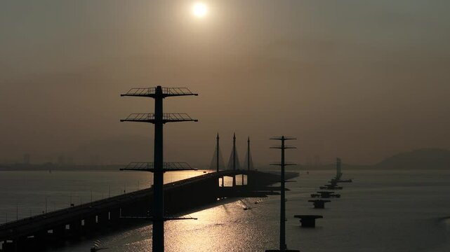 Penang Bridge at sunrise, framed by two TNB monopole electric transmission towers. The golden morning light casts a warm glow over the scene, highlighting the bridge's structure.