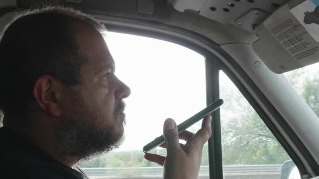 A truck driver with short hair sends a voice message on his smartphone while driving. This dangerous behavior highlights the risks and illegality of distracted driving.