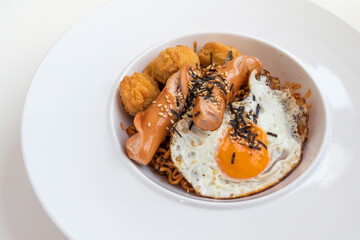 Spicy korea noodle fried egg sausages and fried chicken topped with seaweed and white sesame seeds in a white plate.