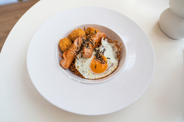 Spicy korea noodle fried egg sausages and fried chicken topped with seaweed and white sesame seeds in a white plate.