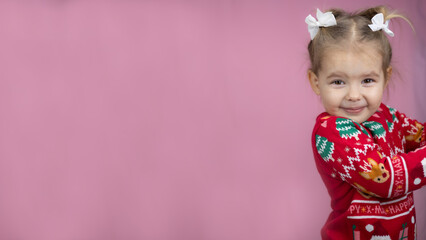 Cute baby girl beams with joy in festive red sweater with deer and Christmas trees. Child radiates joyful festive spirit of new year on bright pink background. New year, christmas, children.