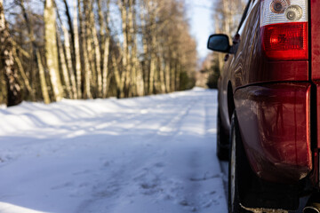 A red car is parked on the snowy side of the road that winds through the forest, and the sun casts...