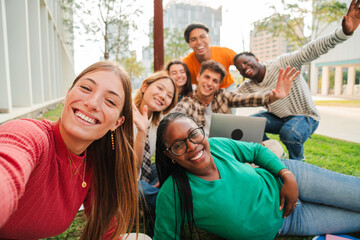 Blonde female student smiling taking a selfie with her classmates sitting at university campus lawn. Big group of teenagers laughing and looking at camera and posing for a portrait at the high school