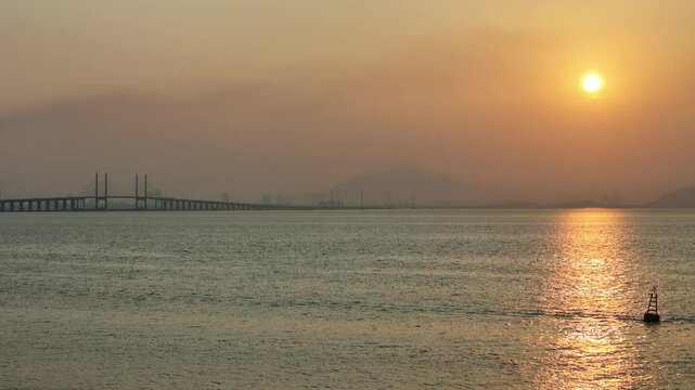Penang Bridge at sunrise, framed by two TNB monopole electric transmission towers. The golden morning light casts a warm glow over the scene, highlighting the bridge's structure.