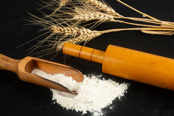 Wooden scoop with flour and rolling pin on black background.