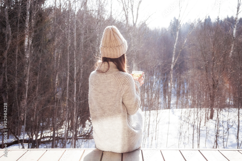 Wall mural Pensive woman looking on winter forest sitting alone drinking hot tea in cold sunny winter day outdoors as loneliness concept