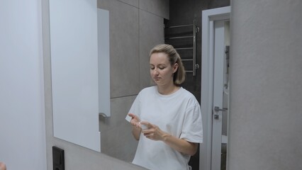 A woman stands in front of a bathroom mirror and applies cream, skin care products and makeup to her face. Young woman applying moisturizer to face as daily cosmetic skin care procedure