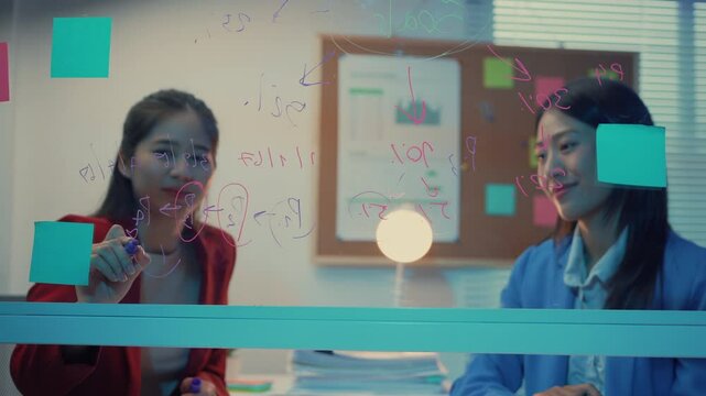 Two businesswomen engage in a strategic discussion within an office setting. Utilizing a glassboard, they brainstorm ideas and collaborate on innovative solutions for their projects