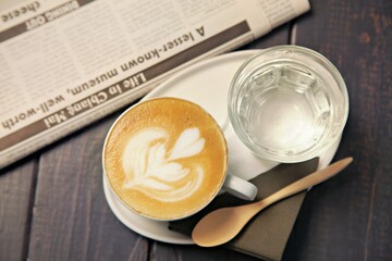 A cozy coffee setup with a latte art, water, and a newspaper on a wooden table.