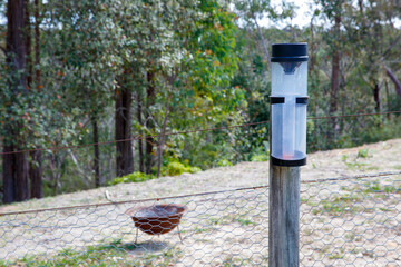 Photograph of a plastic rain gauge on a wooden post near a fence adjacent to bushland on a...