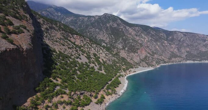 A stunning aerial view of a secluded coastal landscape with rugged mountains and a pristine beach meeting the calm, blue sea. The clear sky and vibrant greenery enhance the natural beauty of this remo